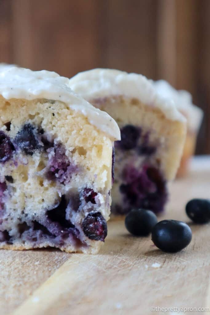 Close up view of a blueberry cottage cheese muffin cut in half on a cutting board. There is cream cheese frosting on the muffin and three blueberries in front.