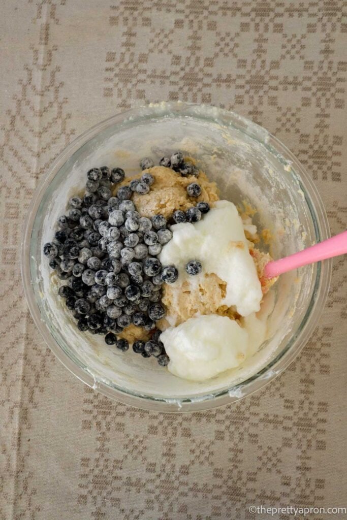 Fresh blueberries and whipped egg whites placed in bowl with beige muffin batter in glass bowl.