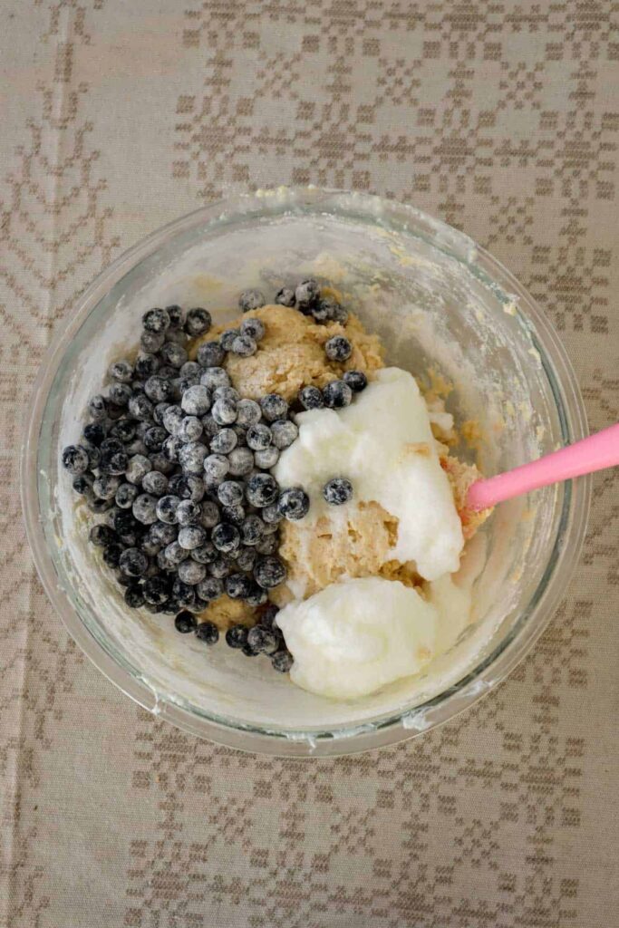 Fresh blueberries and whipped egg whites placed in bowl with beige muffin batter in glass bowl.