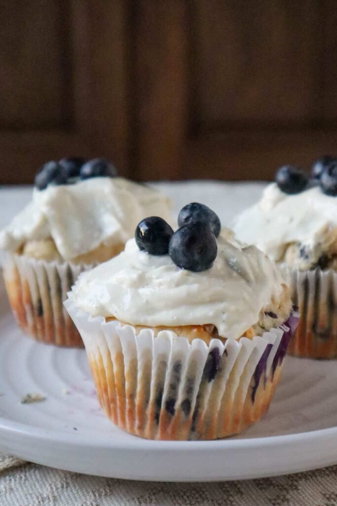 Three blueberry cottage cheese muffins on a plate with white frosting and three blueberries on top of each muffin. There is a wooden cupboard in the background.