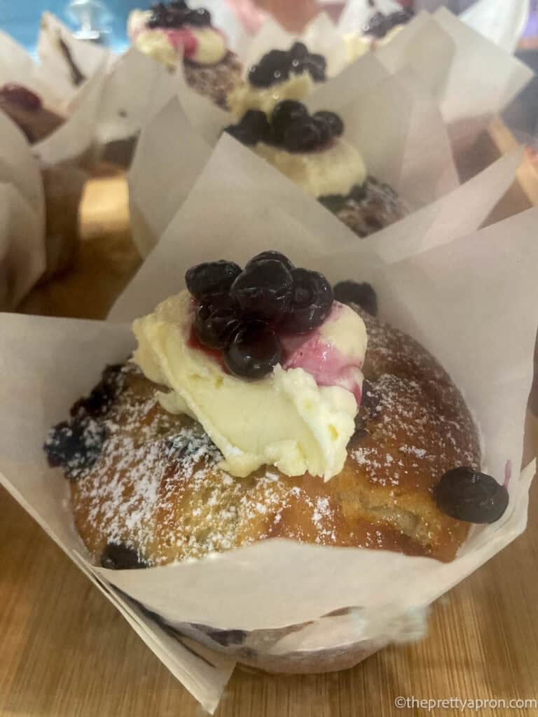 Blueberry muffin with dollop of cream on top with blueberries in syrup on top. The muffin is enveloped with baking paper and is sitting on a shelf. You can see other blueberry muffins in the background.