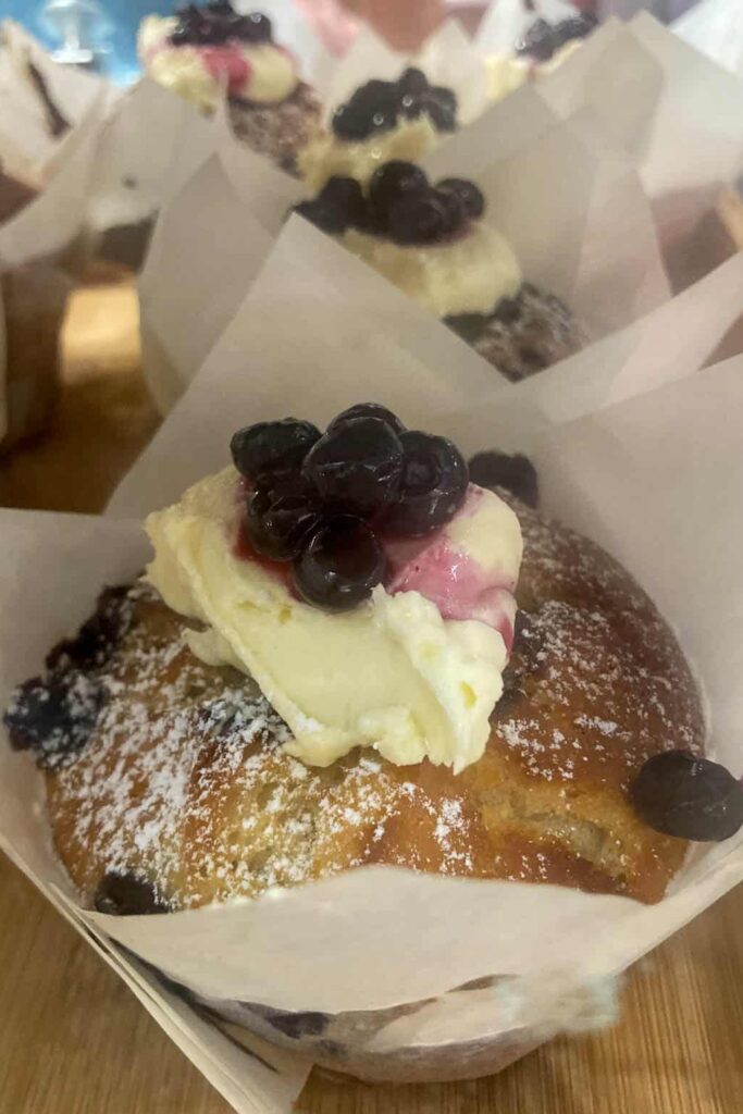 Blueberry muffin with dollop of cream on top with blueberries in syrup on top. The muffin is enveloped with baking paper and is sitting on a shelf. You can see other blueberry muffins in the background.