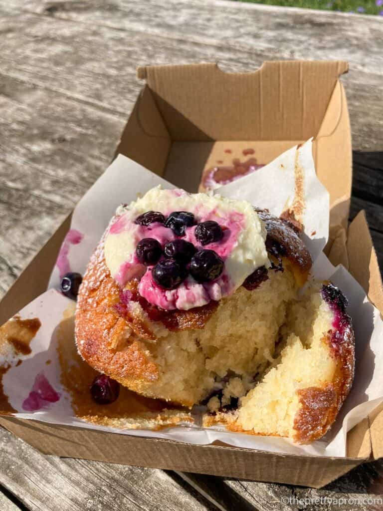 Blueberry muffin with dollop of cream on top with blueberries in syrup on top. The muffin is in a cardboard takeaway box and is lying on a wooden picnic table.