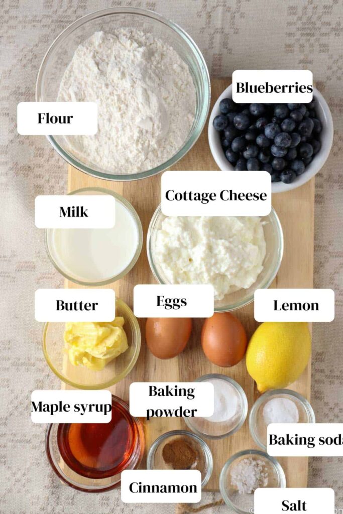 Blueberries, cottage cheese, flour, lemon, butter, two eggs, baking soda in small bowl, baking powder in small bowl, maple syrup in small glass bowl, cinnamon in small glass jar, all on a wooden cutting board, which is on a linen tablecloth.