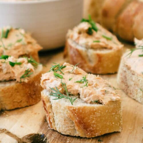 Five slices of baguettes toasted with salmon cream cheese topping and fresh herb garnish. Toasts are served on wooden board and small bowl of salmon cream cheese mixture is seen in background.