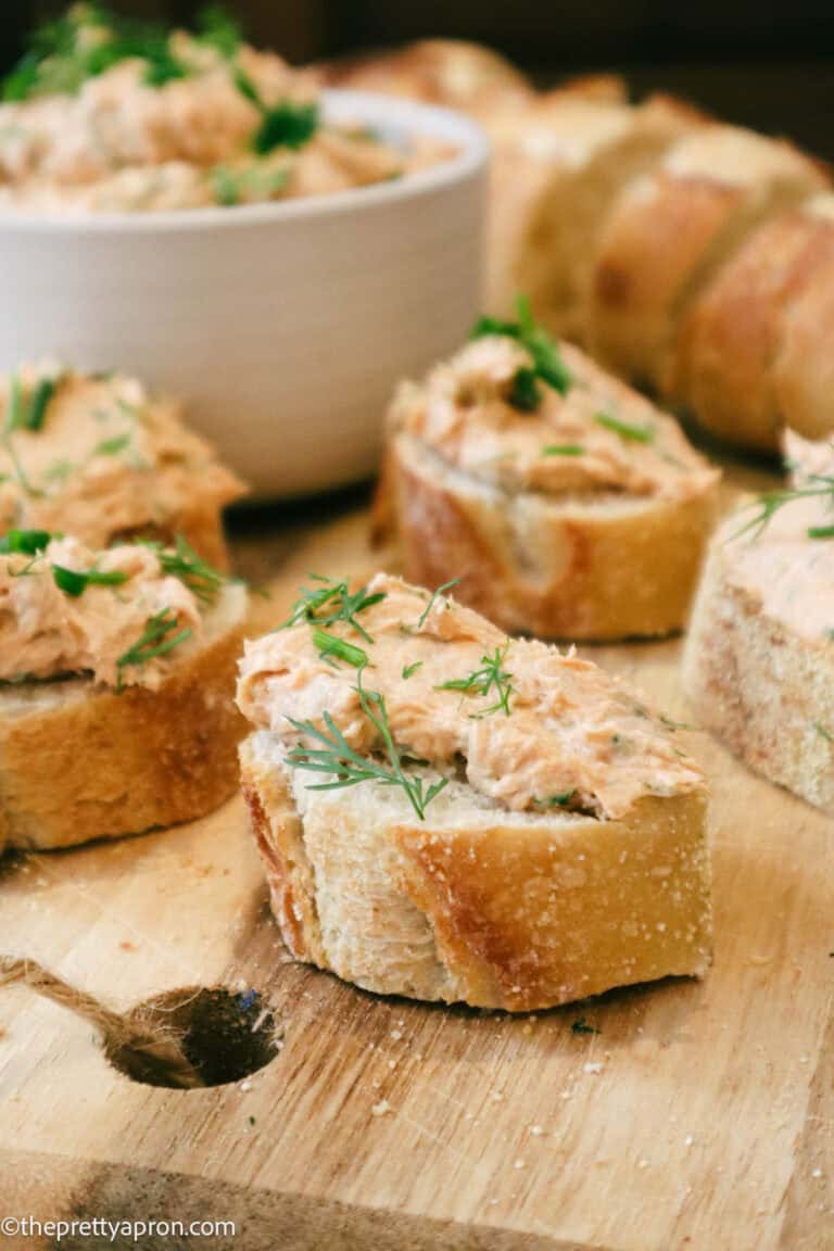 Five slices of baguettes toasted with salmon cream cheese topping and fresh herb garnish. Toasts are served on wooden board and small bowl of salmon cream cheese mixture is seen in background.