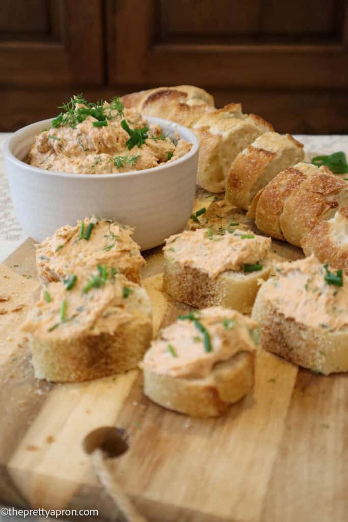 Canned salmon and cream cheese spread in small bowl placed on wooden cutting board, along with slices of sourdough baguette and five salmon cream cheese toasts topped with chopped fresh dill, chives and parsley