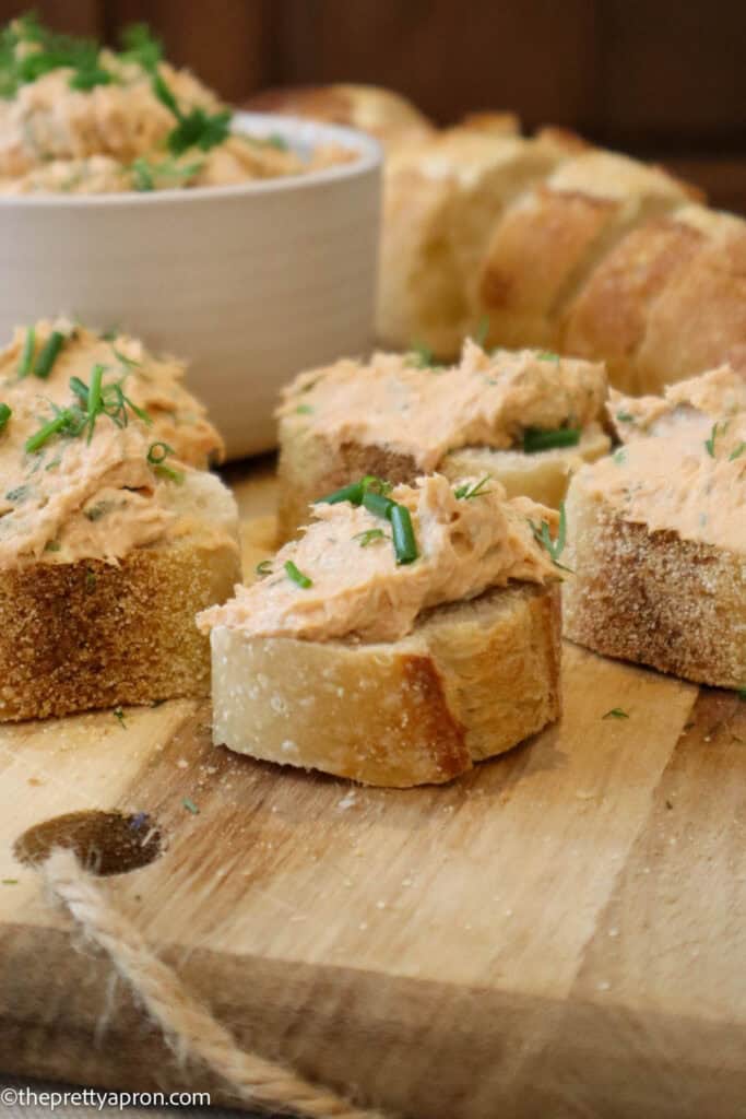 Close up of salmon cream cheese spread on slice of toast. There are 5 toasts on a wooden cutting board and a small bowl of the mixture in the background, along with slices of baguette toasts.