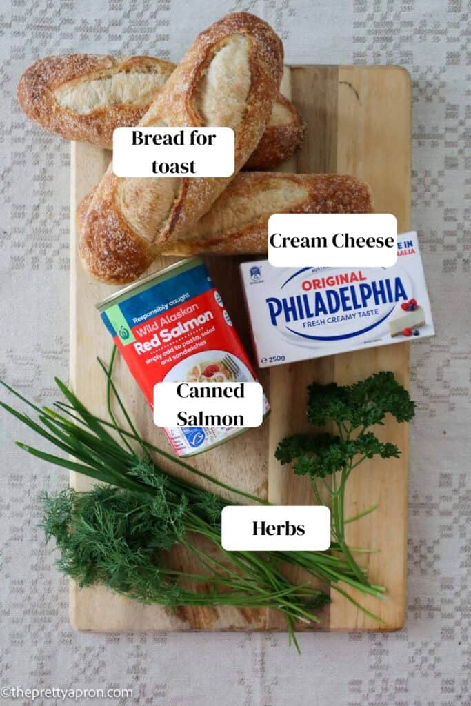 Sourdough bread, canned red salmon, fresh dill, chives, spring onion and parsley and cream cheese alls placed on a wooden chopping board, which is placed on a linen napkin.