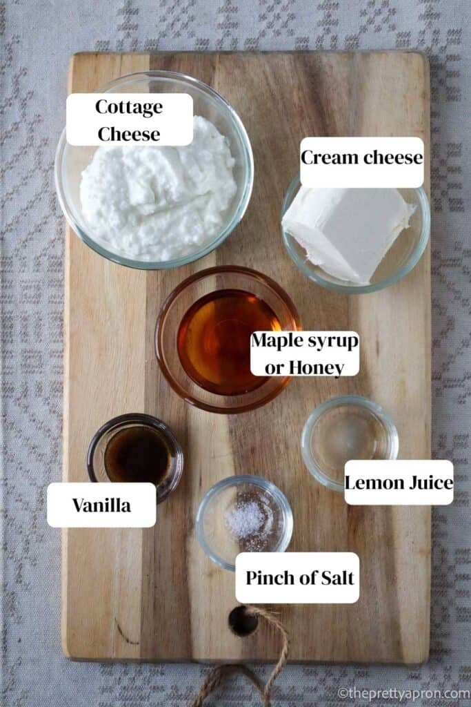 Cottage cheese, cream cheese, maple syrup, lemon juice, vanilla, pinch of salt all in small glass bowl on a wooden cutting board. The board is on a linen beige table cloth.
