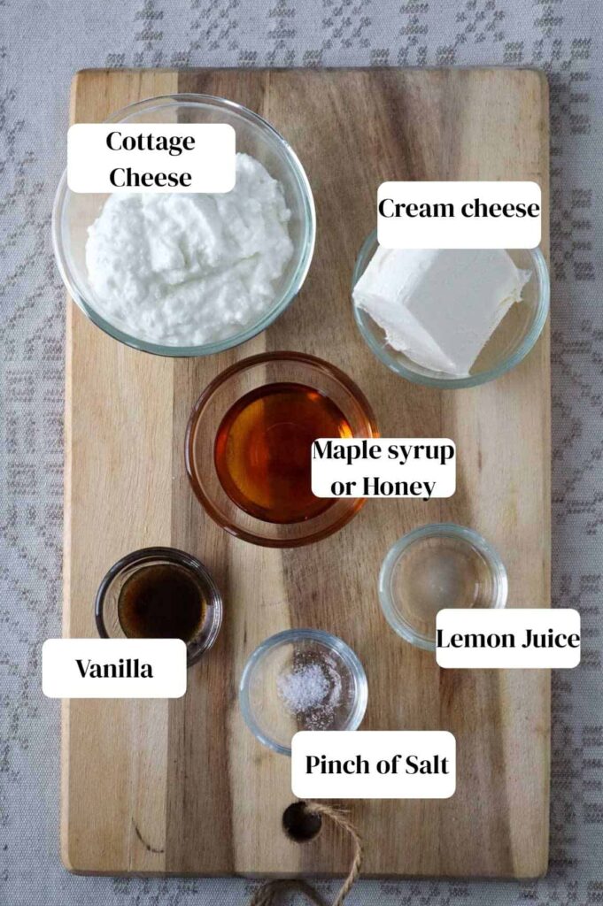 Cottage cheese, cream cheese, maple syrup, lemon juice, vanilla, pinch of salt all in small glass bowl on a wooden cutting board. The board is on a linen beige table cloth.