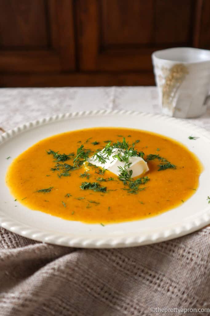 bowl of pumkin cauliflower soup with dollop of sour creamn and chopped dill on linen tablecloth. There is a beige coffee cup in the background.