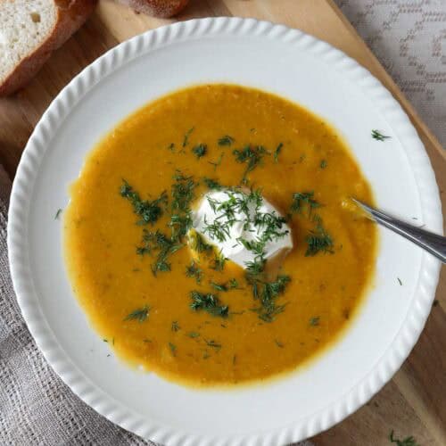 Orange colored pumpkin and cauliflower soup with dollop of sour cream and chopped dill on white bowl on wooden cutting board. there is a beige coffee cup in the background.