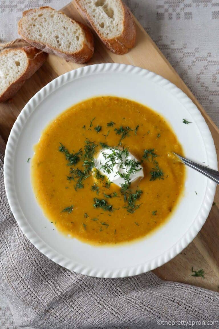 Orange colored pumpkin and cauliflower soup with dollop of sour cream and chopped dill on white bowl on wooden cutting board. there is a beige coffee cup in the background.