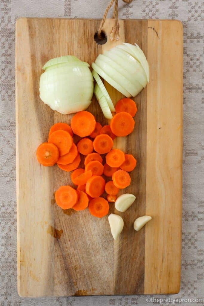 Sliced onion, carrot and three garlic cloves on wooden chopping board.