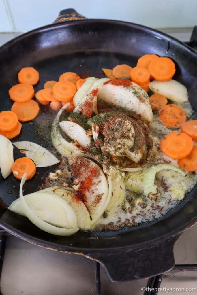 Sliced onions, carrots, garlic, juniper berries, smoked paprika, cinnamon and caraway seeds frying in a black skillet.