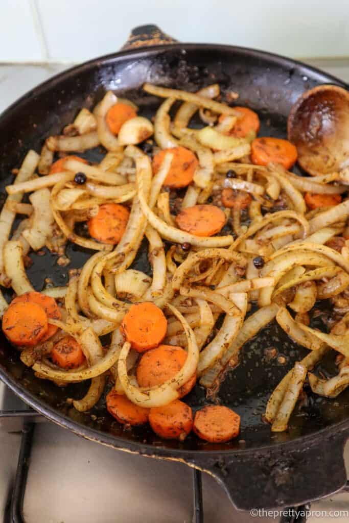 Sliced onions, carrots, garlic, juniper berries, smoked paprika, cinnamon and caraway seeds frying in a black skillet.