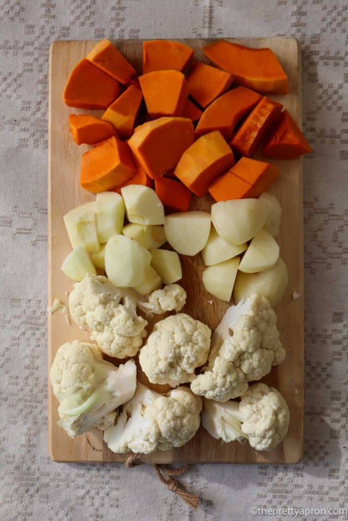 Peeled and chopped pumpkin, potatoes and apple and cauliflower florets on a wooden chopping board.
