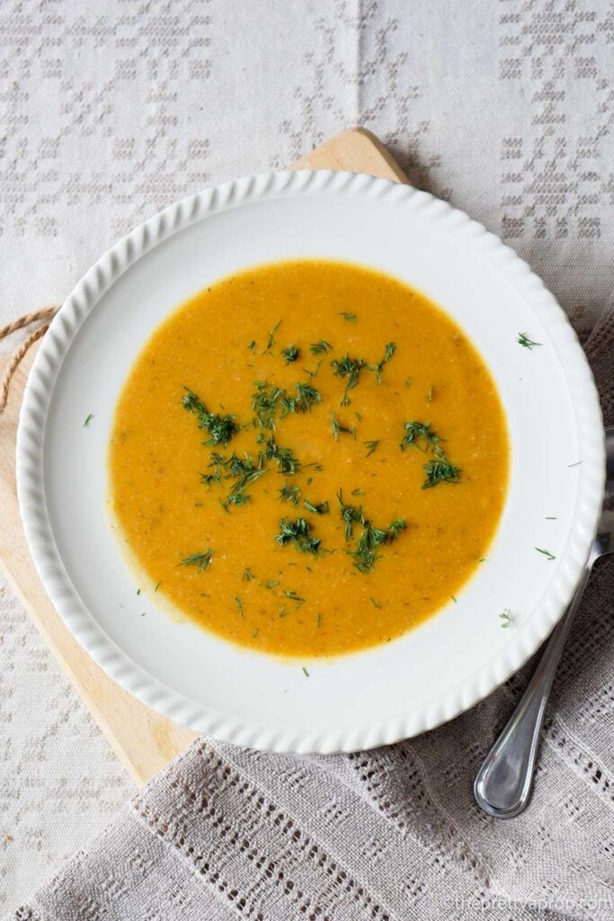 bowl of orange pumpkin cauliflower soup i a white bowl on a wooden board, which is set on a linen tablecloth. The soup is sprinkled with green dill.