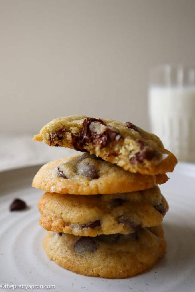 stack of chocolate chip cookies with top cookie already bitten into and glass of milk in the background