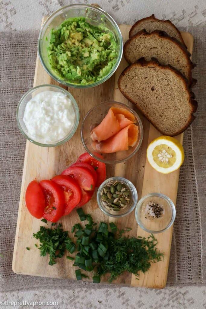 smashed avocado in bowl, pieces of toast, sliced tomato, slices of smoked salmon, cottage cheese in bowl, chopped fresh herbs, pumpkin and sunflower seeds in small bowl, salt and pepper, all on a wooden cutting board