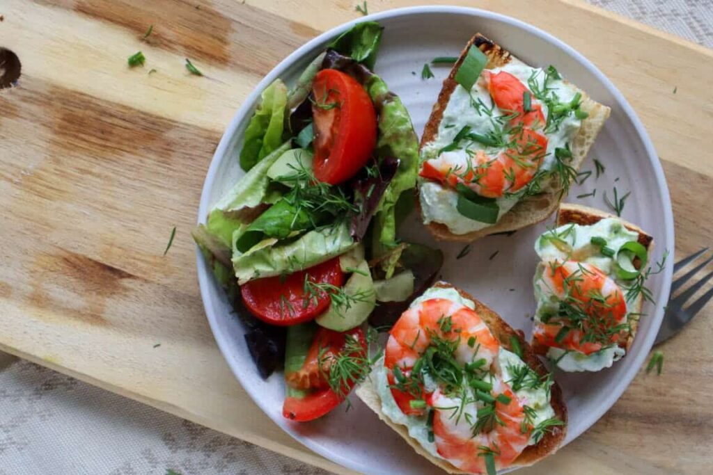 shrimp avocado toast on plate with garden salad and plate on wooden board