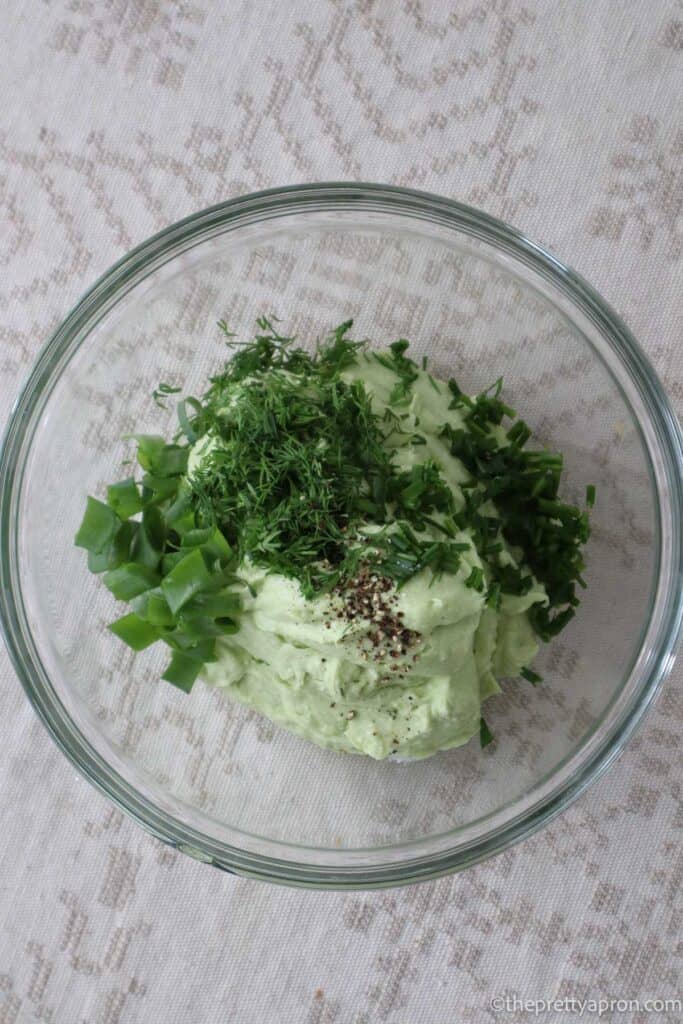 Light green creamy dip mixture in a glass bowl topped with spoonfuls of chopped dill, parsley and chives and some salt and pepper.