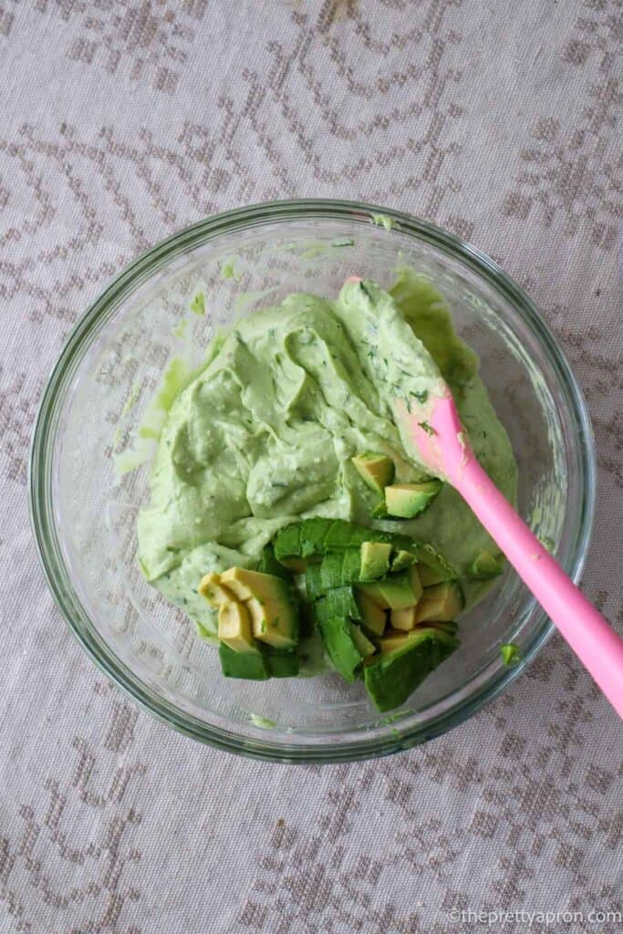 Green avocado cottage cheese dip mixture in glass bowl with half and avocado chopped in small pieces on top. There is a pink spatula that is mixing it.