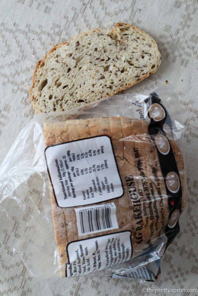 Slice of sourdough bread next to Vital Rye loaf of bread in a plastic bag. This bag is on a linen tablecloth.