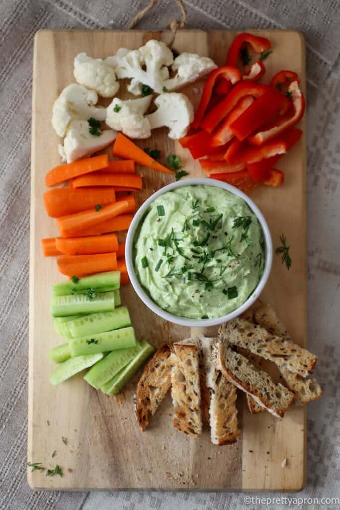 Green avocado and cottage cheese dip on wooden cutting board surrounded by bell pepper, cauliflower, carrot, cucumber and bread sticks.
