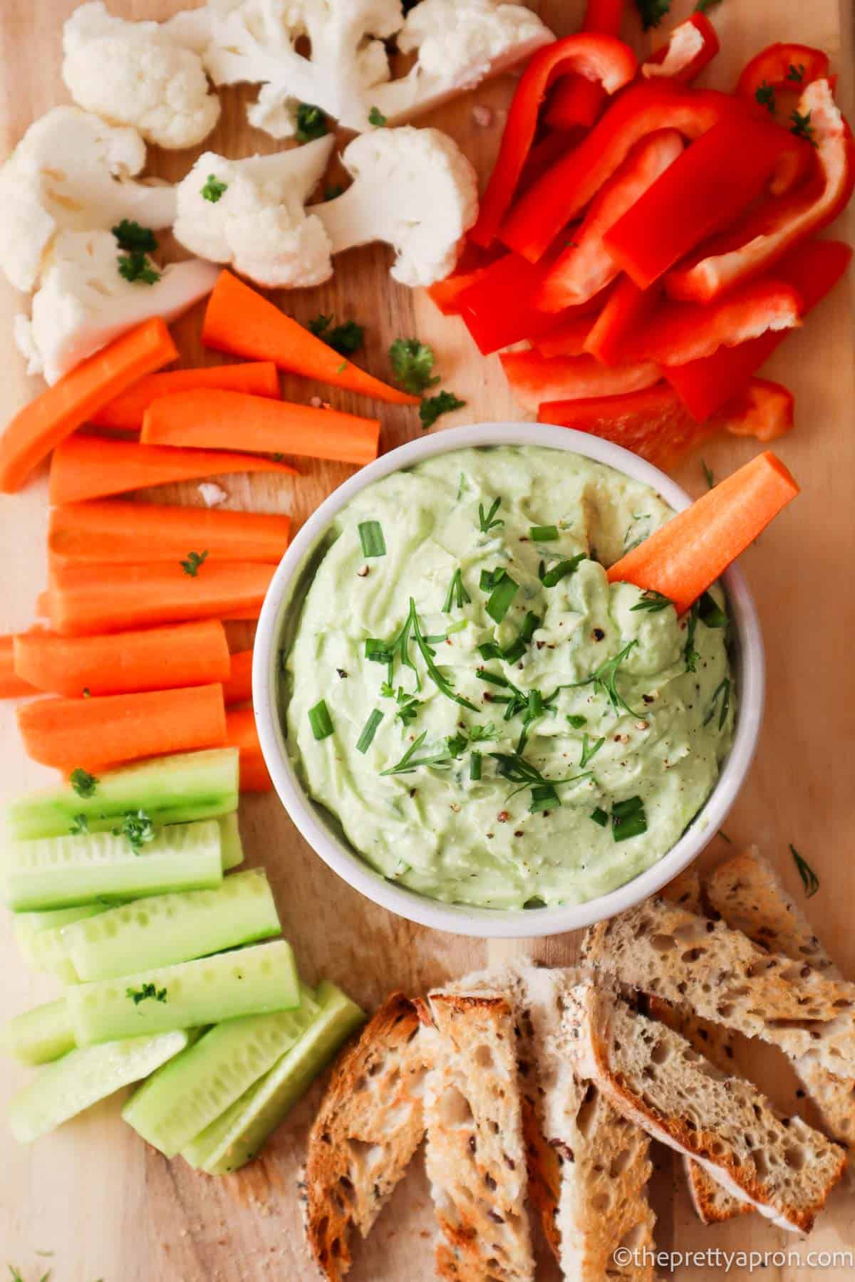 Green avocado and cottage cheese sip in a small white bowl. There is a carrot slice dipped in the bowl. there are carrot sticks, red pepper, cucumber, cauliflower and bread sticks surrounding the bowl and a wooden board.