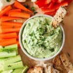 Green avocado cottage cheese dip in small white bowl with thin slice of toasted bread dipped in it. There are carrot, cauliflower, red pepper and cucumber sticks around the bowl.