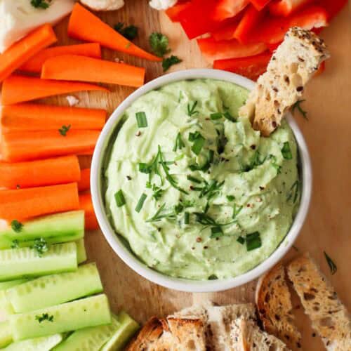 Green avocado cottage cheese dip in small white bowl with thin slice of toasted bread dipped in it. There are carrot, cauliflower, red pepper and cucumber sticks around the bowl.