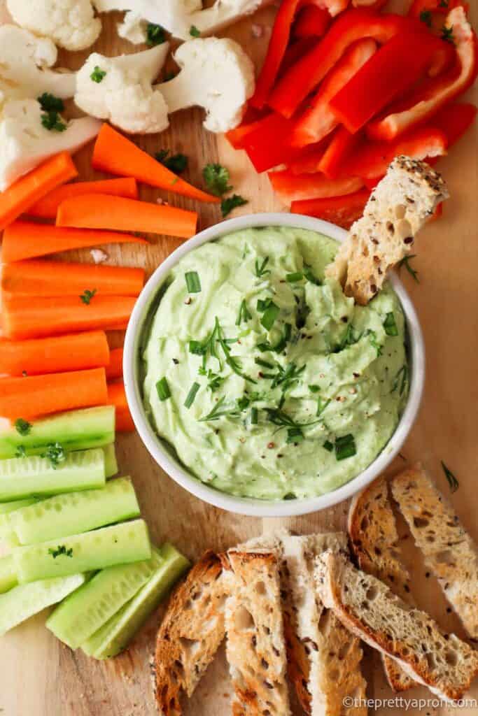 Green avocado cottage cheese dip in small white bowl with thin slice of toasted bread dipped in it. There are carrot, cauliflower, red pepper and cucumber sticks around the bowl.