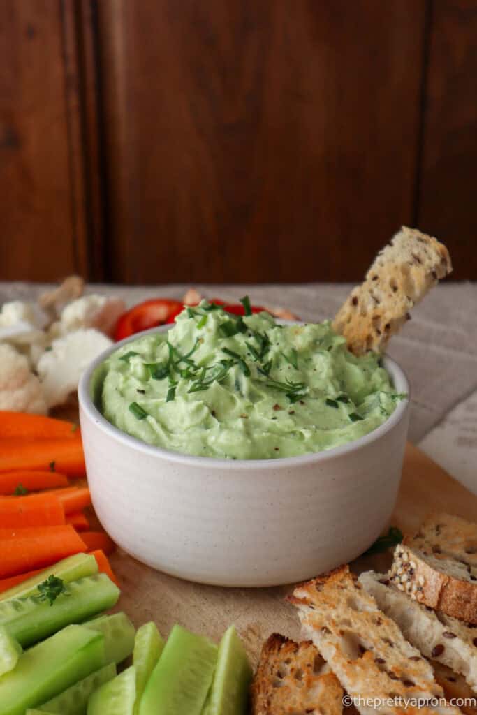 Green avocado cottage cheese dip in small white bowl with thin slice of toasted bread dipped in it. there are carrot, cauliflower, red pepper and cucumber sticks around the bowl. There is a wooden cupboard in the background.