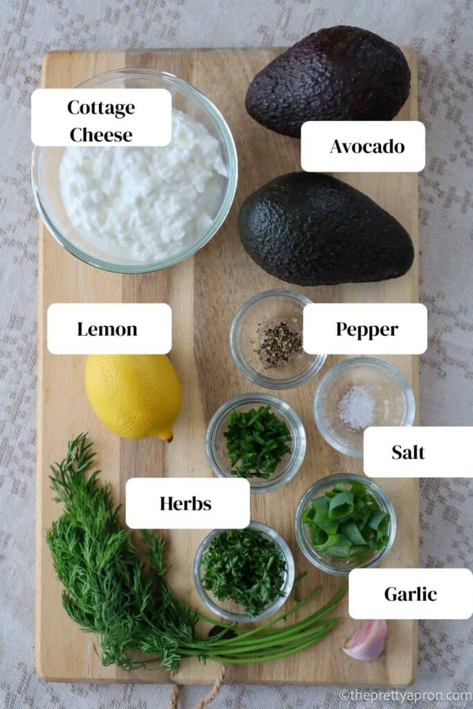 two avocados, cottage cheese in small glass bowl, lemon, parsley, chives and spring onion each chopped up in a very small glass bowl, bunch of dill, salt and pepper each in a small glass bowl, all sitting on a wooden cutting board.