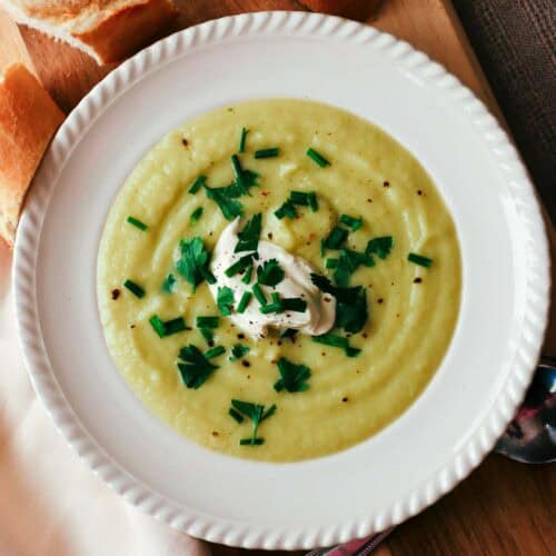 Cauliflower, potato and leek soup with sprinkles of chives and parsley and a dollop of creme fraiche in an off-white bowl. The bowl is on a wooden cutting board and there are three baguette slices next to it.