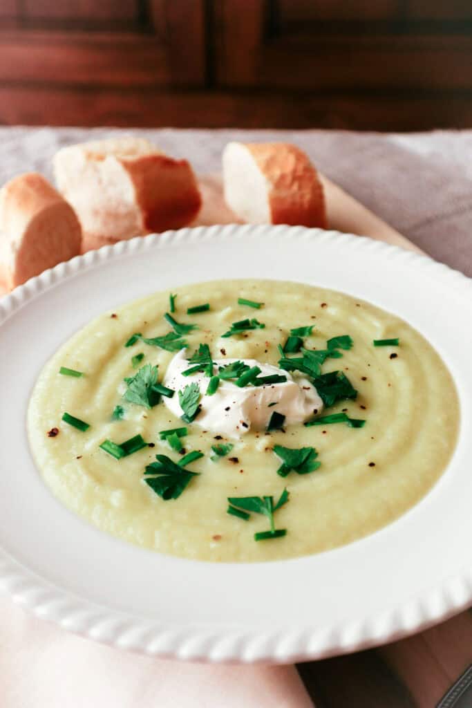 cauliflower, potato and leek soup in white bowl with parsley garnish and three slices of baguette nearby. All on a linen tablecloth with wooden kitchen cupboard in background.