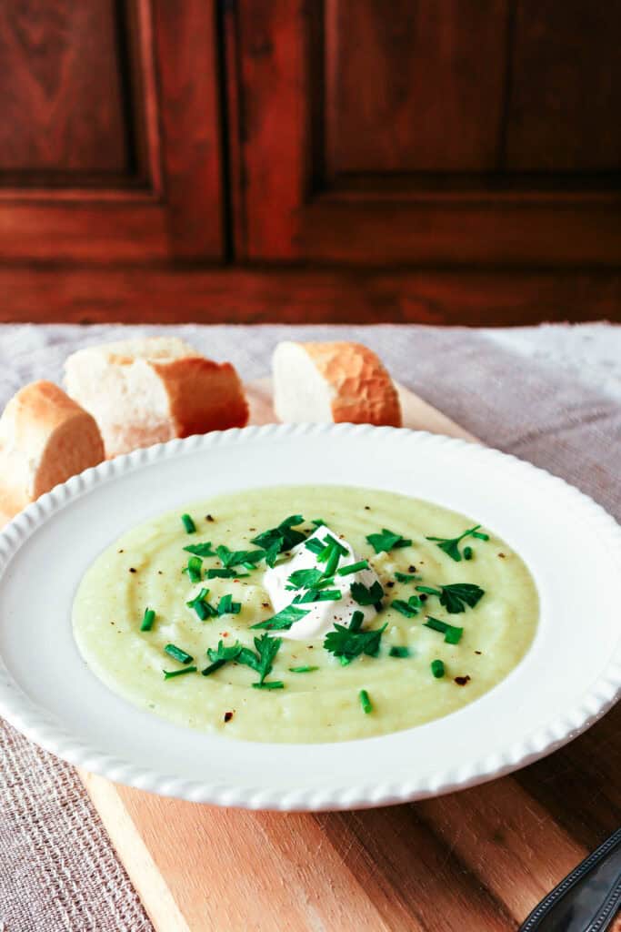 cauliflower, potato and leek soup in white bowl with parsley garnish and three slices of baguette nearby. All on a linen tablecloth with wooden kitchen cupboard in background.
