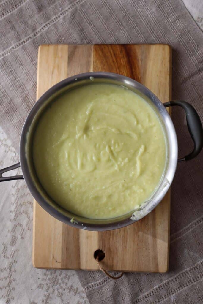 Blended cauliflower, leek and potato soup in a stainless steel pot on a wooden cutting board.