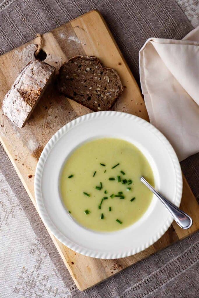 Cauliflower, leek and potato soup in off white bowl. There are spinkles of chives. Thebowl is on a wooden cutting board, next to a loaf and slice of rustic rye bread.