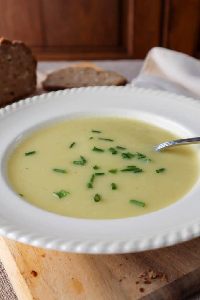 Off-white bowl of light yellow soup with chives with a spoon in it. The bowl is on a wooden cutting board and there is a slice of dark rye bread in the background.