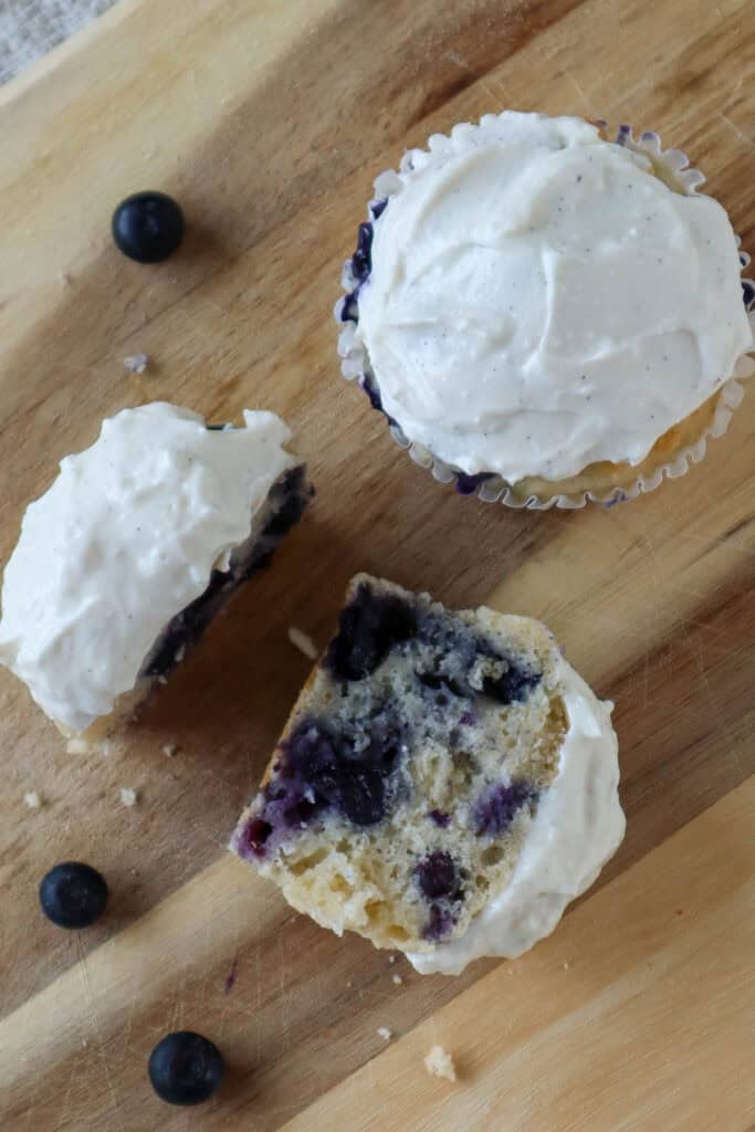 two blueberry cottage cheese muffins with cottage cheese frosting on a wooden board. One of the muffins is cut in half.