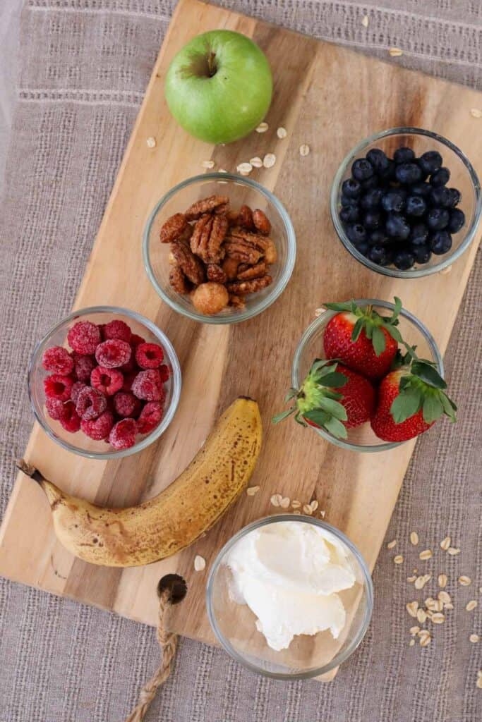Strawberries, blueberries, frozen raspberies, a banana, maple pecans, yogurt and an apple on a wooden board.