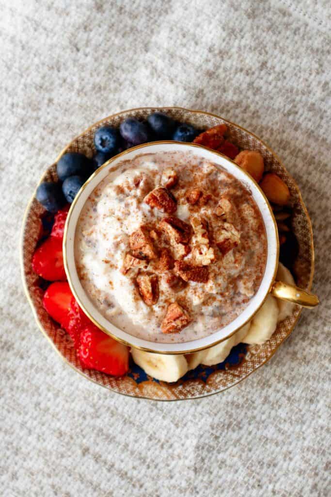 Cinnamon roll overnight oats served in a pretty teacup surrounded by strawberries, bananas, blueberries and nuts in the saucer.