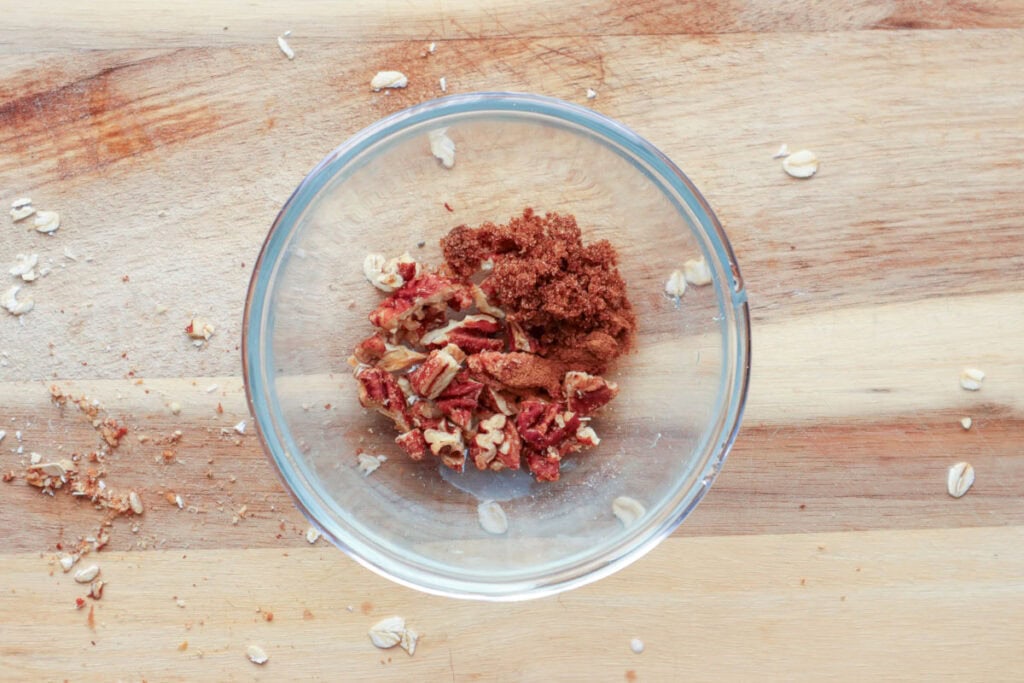 Cinnamon, brown sugar and nuts in small glass bowl on wooden board.