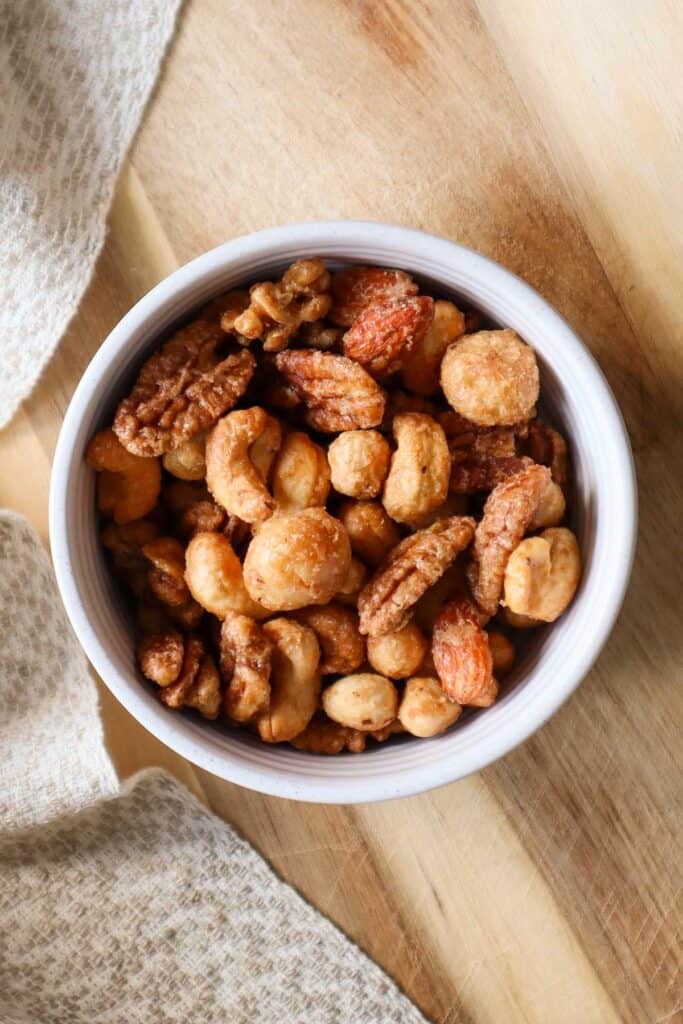 roasted maple nuts in a beige bowl on a wooden board. Roasted nuts recipe includes maple roasted pecans, maple roasted walnuts, maple roasted macadamia nuts, maple roasted hazelnuts and maple roasted almonds.