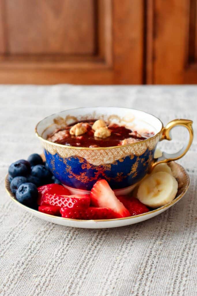 Oats with nutella sauce and hazelnuts in a teacup with blueberries, strawberries and bananas in saucer. Teacup and saucer are placed in linen tablecloth.