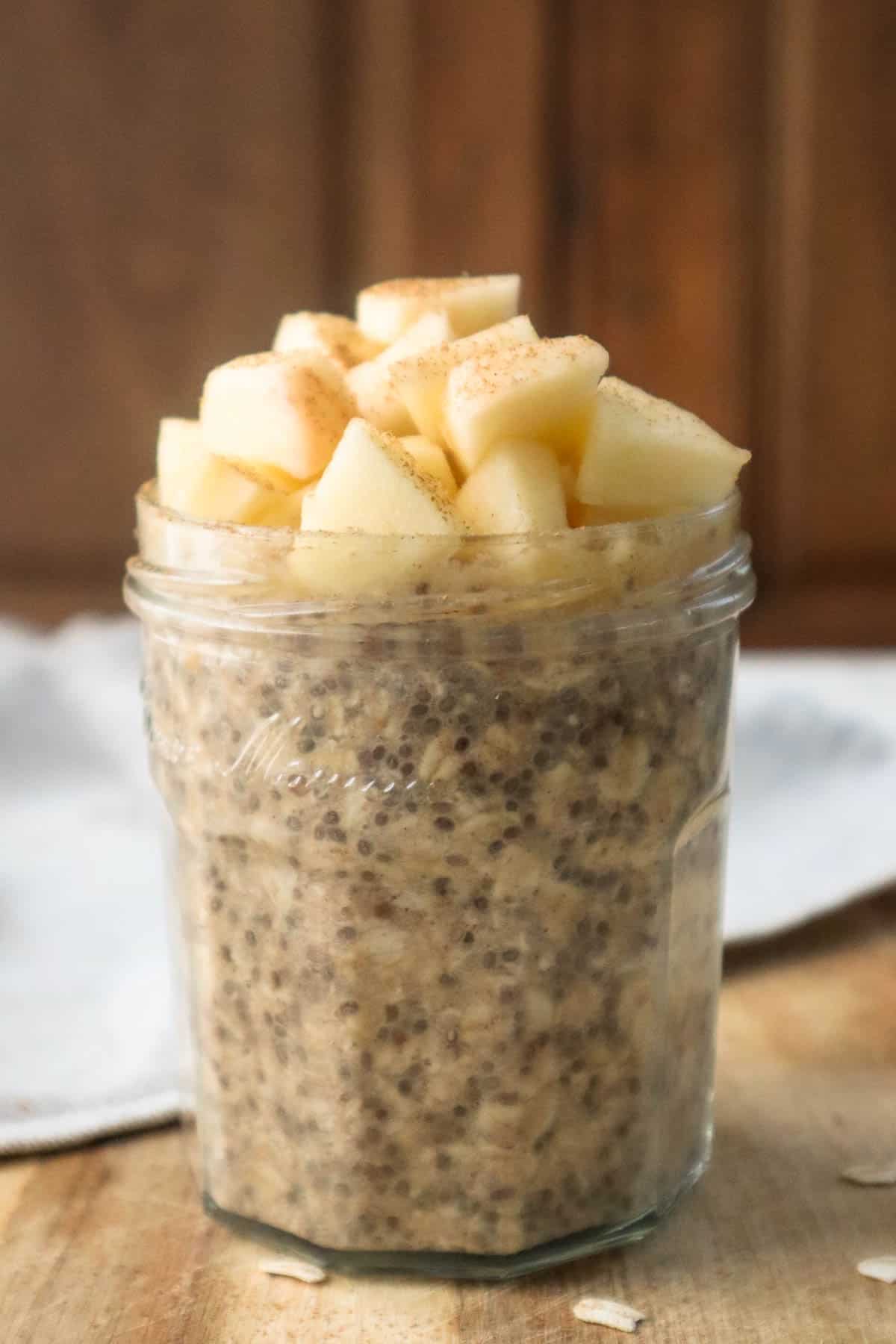 Applesauce overnight oats in glass jar topped with chopped apples on wooden board and beige napkin next to it.
