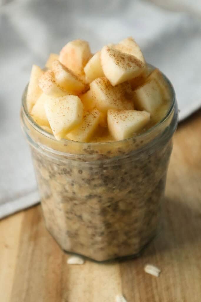 Applesauce overnight oats topped with chopped apples in a glass jam jar that is sitting on a wooden cutting board.